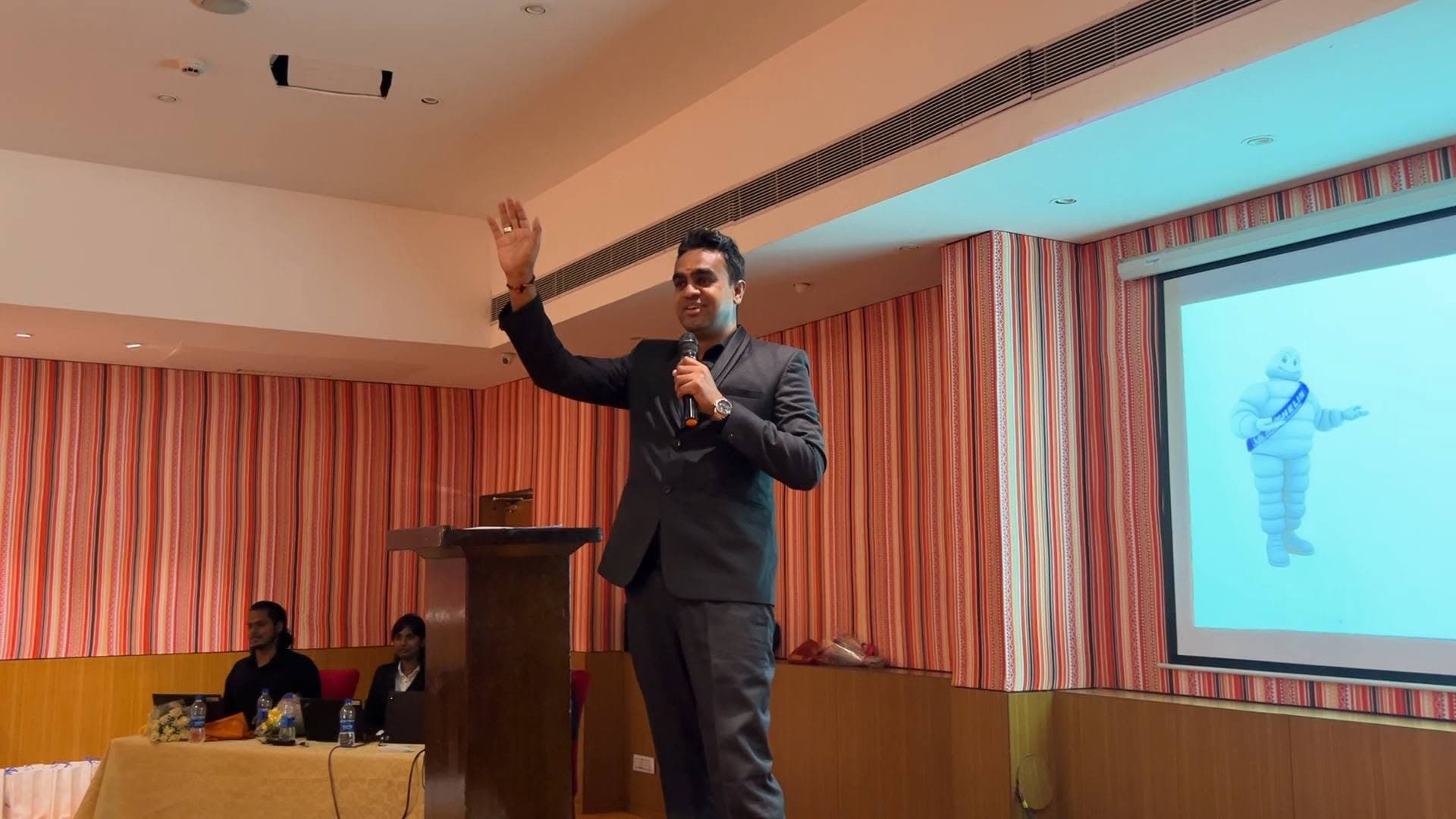 Manesh in a dark suit waving to the audience from stage